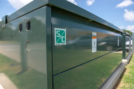 Closeup of one corner of S&C’s Vista Underground Distribution Switchgear. The green S&C logo is in the foreground. In the background is a blue sky with puffy white clouds.